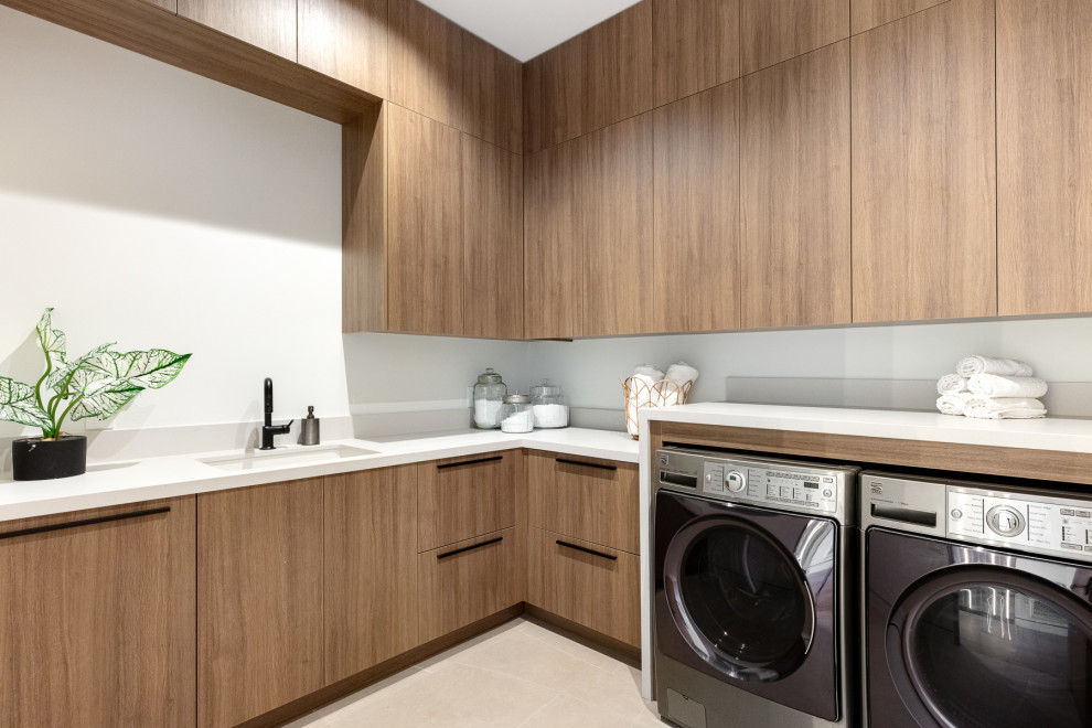 Photo of a contemporary l-shaped laundry room in Los Angeles with an undermount sink, flat-panel cabinets, medium wood cabinets, marble benchtops, white splashback, white walls, a side-by-side washer and dryer, beige floor and white benchtop.
