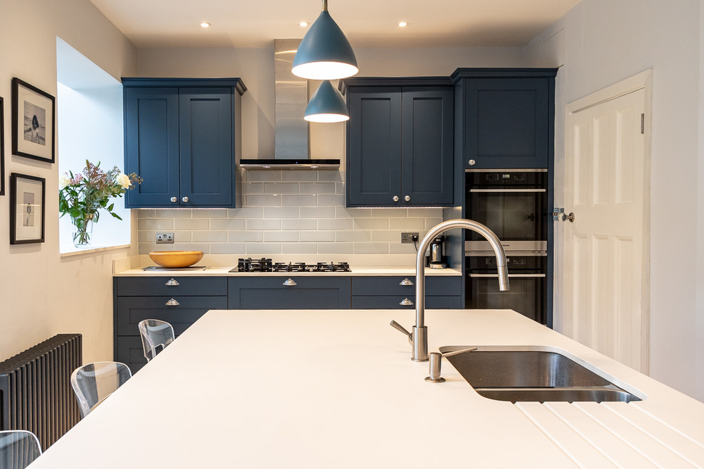 Mid-sized contemporary l-shaped open plan kitchen in London with an integrated sink, shaker cabinets, blue cabinets, solid surface benchtops, stainless steel appliances, light hardwood floors, with island, beige floor and white benchtop.