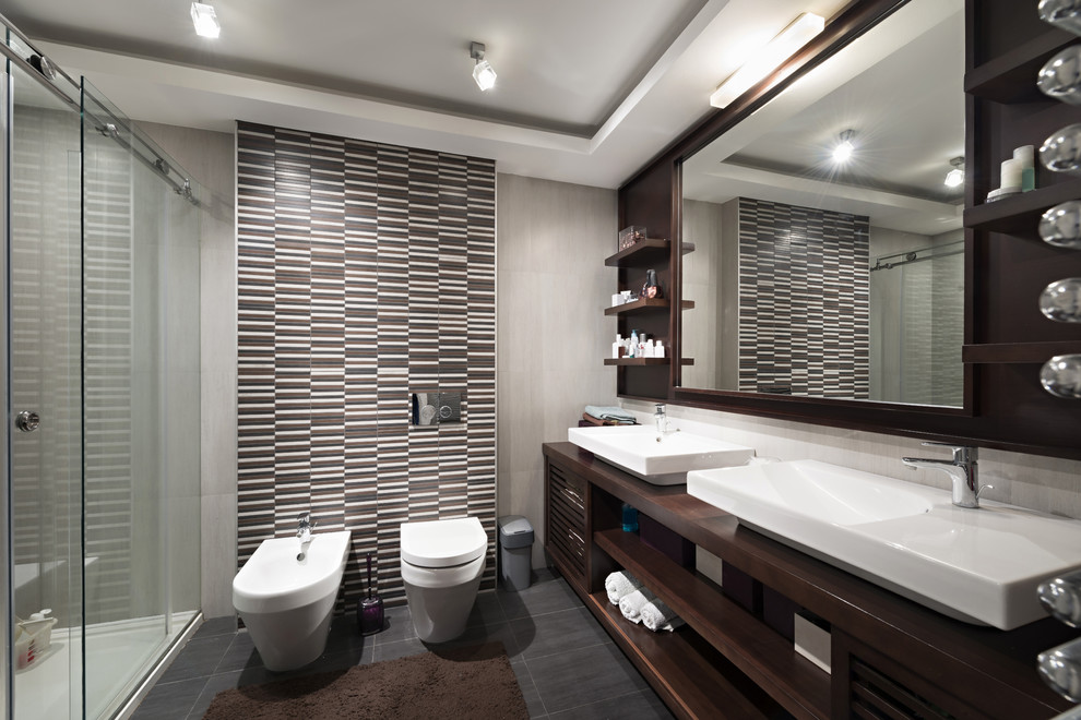 This is an example of a mid-sized contemporary master bathroom in Los Angeles with furniture-like cabinets, dark wood cabinets, a double shower, a bidet, multi-coloured tile, matchstick tile, grey walls, ceramic floors, an integrated sink, wood benchtops, grey floor and a sliding shower screen.
