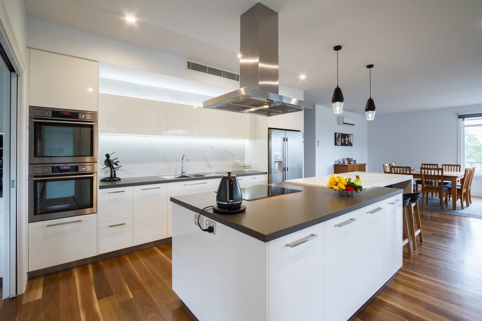 Inspiration for a large modern galley open plan kitchen in Melbourne with a drop-in sink, flat-panel cabinets, white cabinets, quartz benchtops, stone slab splashback, stainless steel appliances, medium hardwood floors and with island.