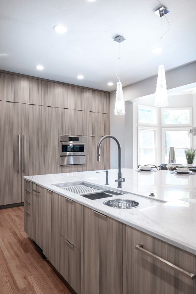 Open concept kitchen - large transitional medium tone wood floor and brown floor open concept kitchen idea with an undermount sink, flat-panel cabinets, brown cabinets, quartzite countertops, white backsplash, ceramic backsplash, stainless steel appliances, an island and beige countertops