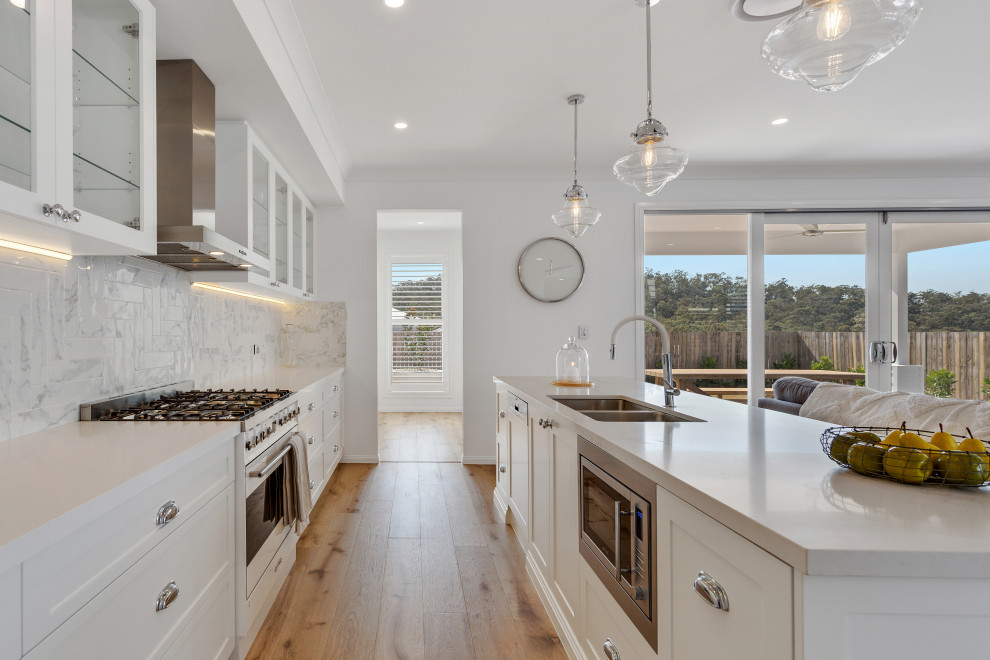Design ideas for an expansive beach style galley open plan kitchen in Brisbane with an undermount sink, glass-front cabinets, white cabinets, quartz benchtops, multi-coloured splashback, ceramic splashback, white appliances, light hardwood floors, with island, multi-coloured floor and white benchtop.