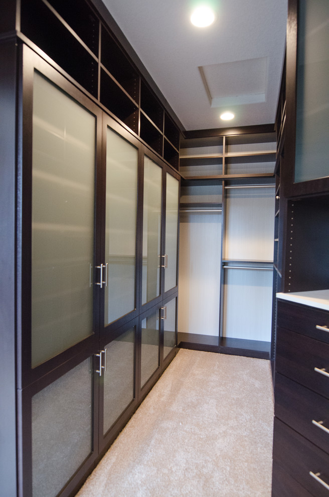 Large gender-neutral walk-in wardrobe in Portland with glass-front cabinets, dark wood cabinets and carpet.