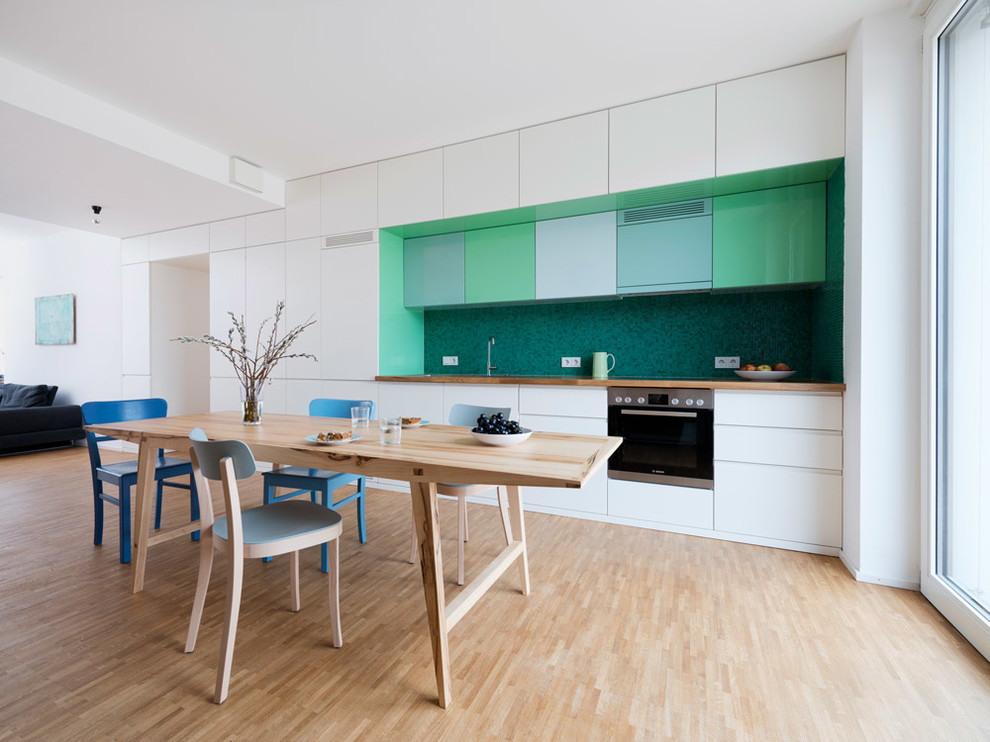 Large scandinavian single-wall eat-in kitchen in Munich with flat-panel cabinets, white cabinets, wood benchtops, green splashback, light hardwood floors and stainless steel appliances.