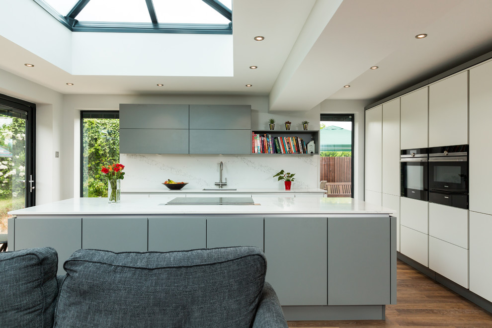 Design ideas for a large contemporary single-wall eat-in kitchen in Hampshire with quartz benchtops, stainless steel appliances, with island, an integrated sink, flat-panel cabinets, grey cabinets, white splashback, stone slab splashback and medium hardwood floors.