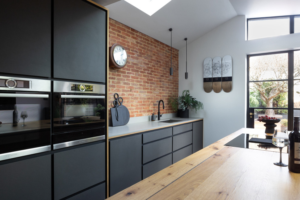 Inspiration for a large contemporary single-wall open plan kitchen in Sussex with a drop-in sink, flat-panel cabinets, distressed cabinets, quartzite benchtops, multi-coloured splashback, brick splashback, black appliances, ceramic floors, with island, grey floor, grey benchtop and vaulted.