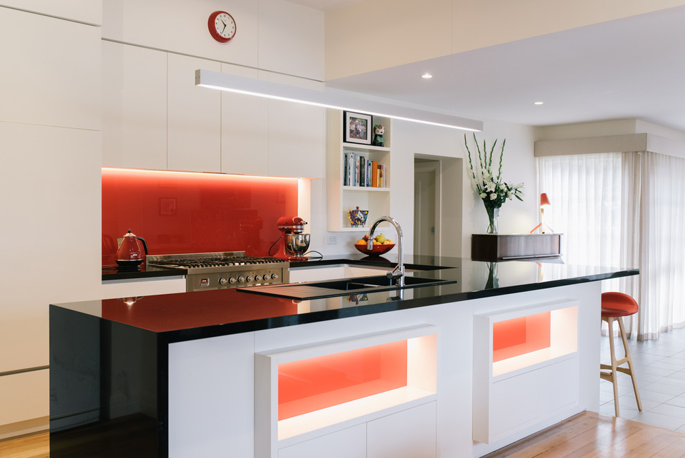 Mid-sized contemporary u-shaped open plan kitchen in Melbourne with a double-bowl sink, flat-panel cabinets, white cabinets, granite benchtops, red splashback, glass sheet splashback, stainless steel appliances, medium hardwood floors and no island.