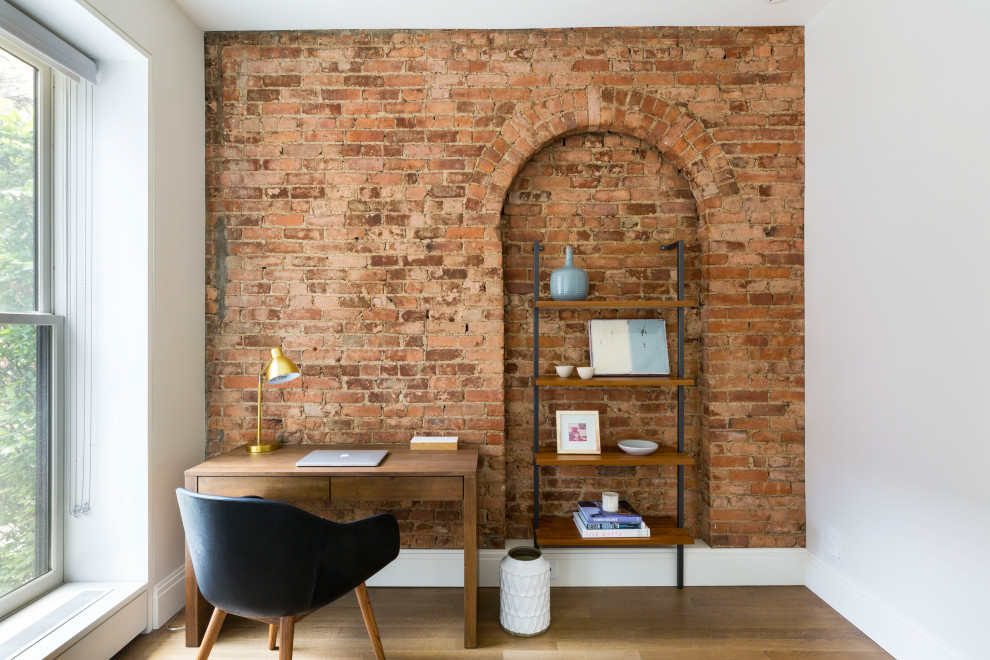 Inspiration for a contemporary home office in New York with white walls, light hardwood floors, a freestanding desk, brown floor and brick walls.
