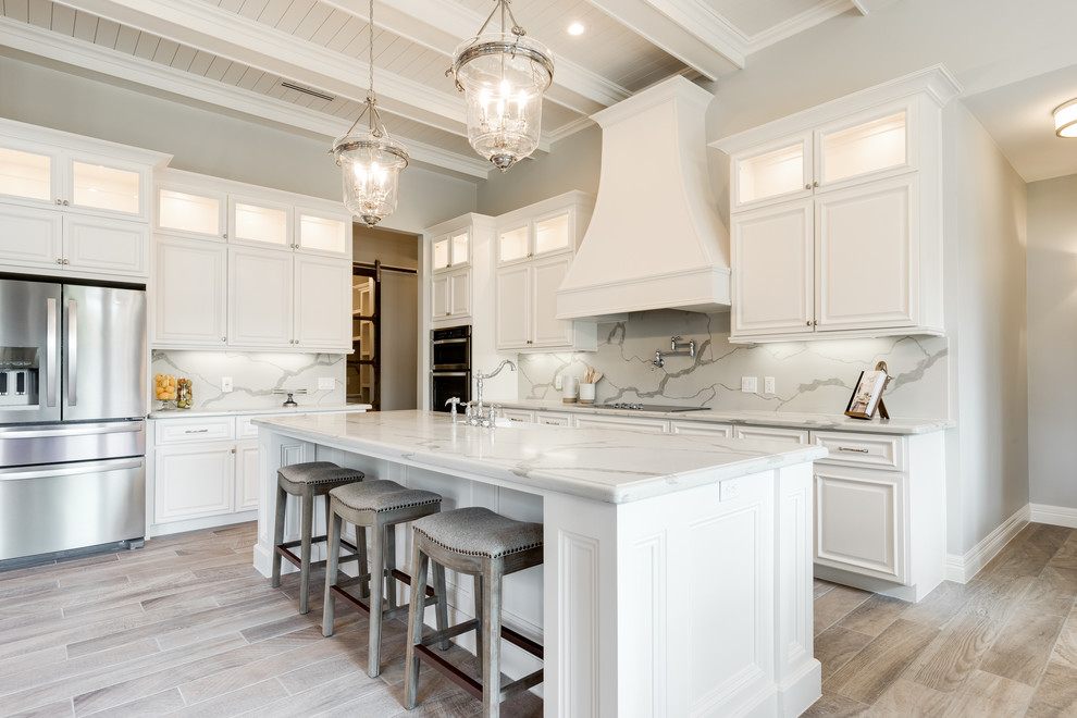 Inspiration for a l-shaped kitchen in Austin with raised-panel cabinets, white cabinets, white splashback, stainless steel appliances, light hardwood floors, with island, beige floor and white benchtop.