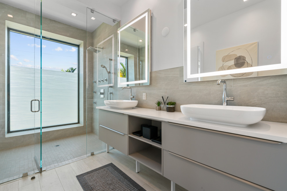 This is an example of an expansive modern master bathroom in Miami with flat-panel cabinets, grey cabinets, a curbless shower, a one-piece toilet, beige tile, porcelain tile, white walls, porcelain floors, a vessel sink, solid surface benchtops, beige floor, white benchtops, a shower seat, a double vanity and a freestanding vanity.