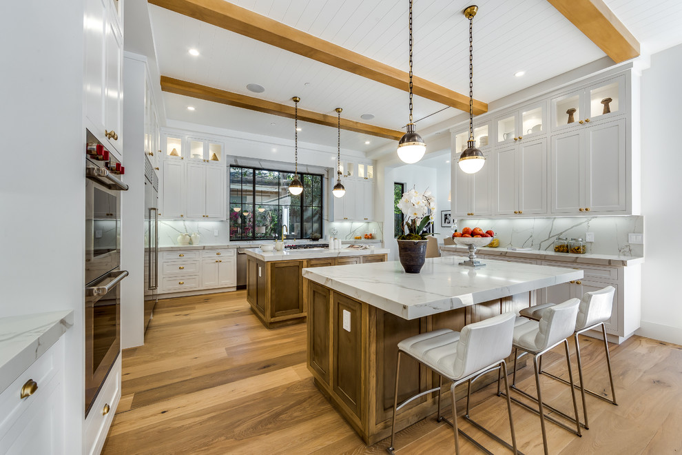 Photo of a mid-sized transitional u-shaped eat-in kitchen in Los Angeles with shaker cabinets, white cabinets, white splashback, stainless steel appliances, medium hardwood floors, multiple islands, a drop-in sink, marble benchtops, brown floor, white benchtop and glass tile splashback.