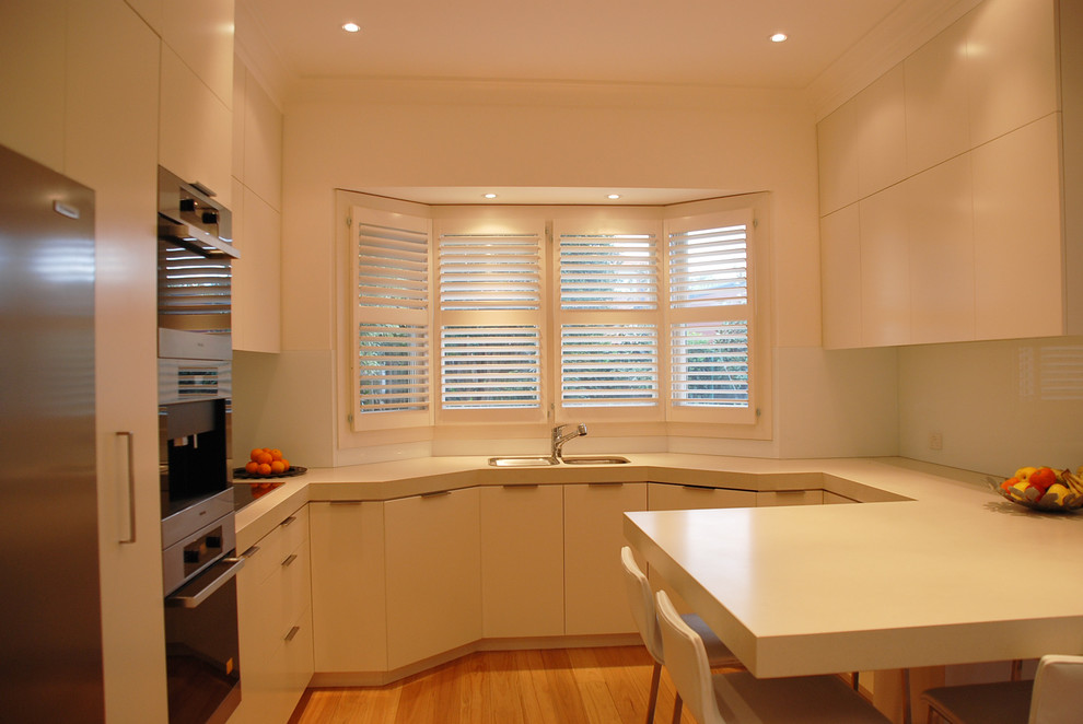Mid-sized contemporary u-shaped eat-in kitchen in Sydney with a double-bowl sink, flat-panel cabinets, white cabinets, quartz benchtops, white splashback, glass sheet splashback, stainless steel appliances, light hardwood floors and a peninsula.