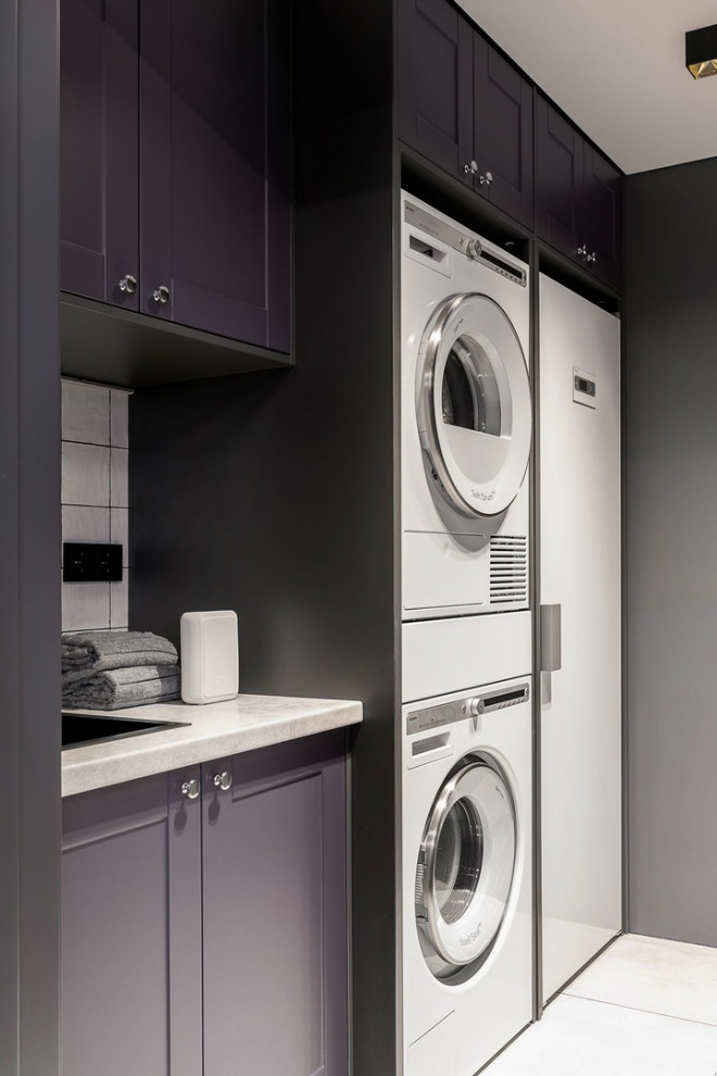 Design ideas for a large contemporary single-wall dedicated laundry room in Other with a drop-in sink, raised-panel cabinets, grey cabinets, laminate benchtops, white splashback, ceramic splashback, grey walls, ceramic floors, a stacked washer and dryer, white floor, white benchtop and panelled walls.