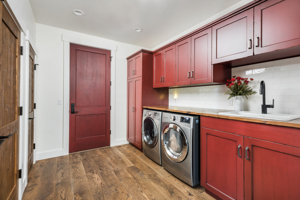 Inspiration for a country single-wall utility room in Boise with a drop-in sink, wood benchtops, white splashback, ceramic splashback, white walls, medium hardwood floors, a side-by-side washer and dryer, brown floor, brown benchtop, shaker cabinets and red cabinets.