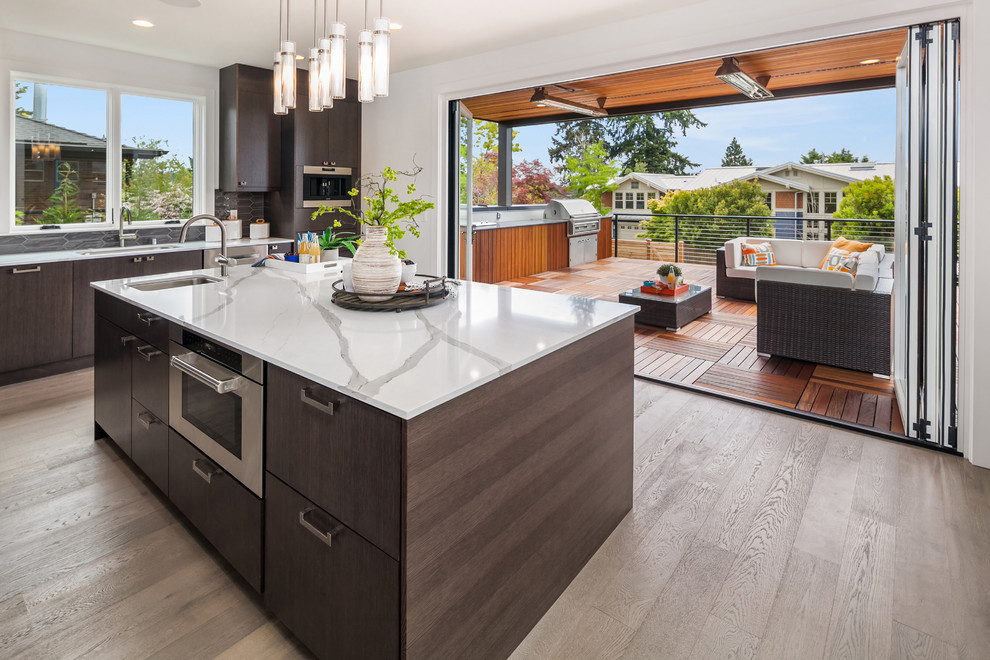 Photo of a large midcentury l-shaped open plan kitchen in Seattle with a drop-in sink, flat-panel cabinets, dark wood cabinets, marble benchtops, grey splashback, ceramic splashback, stainless steel appliances, light hardwood floors, with island, beige floor and white benchtop.