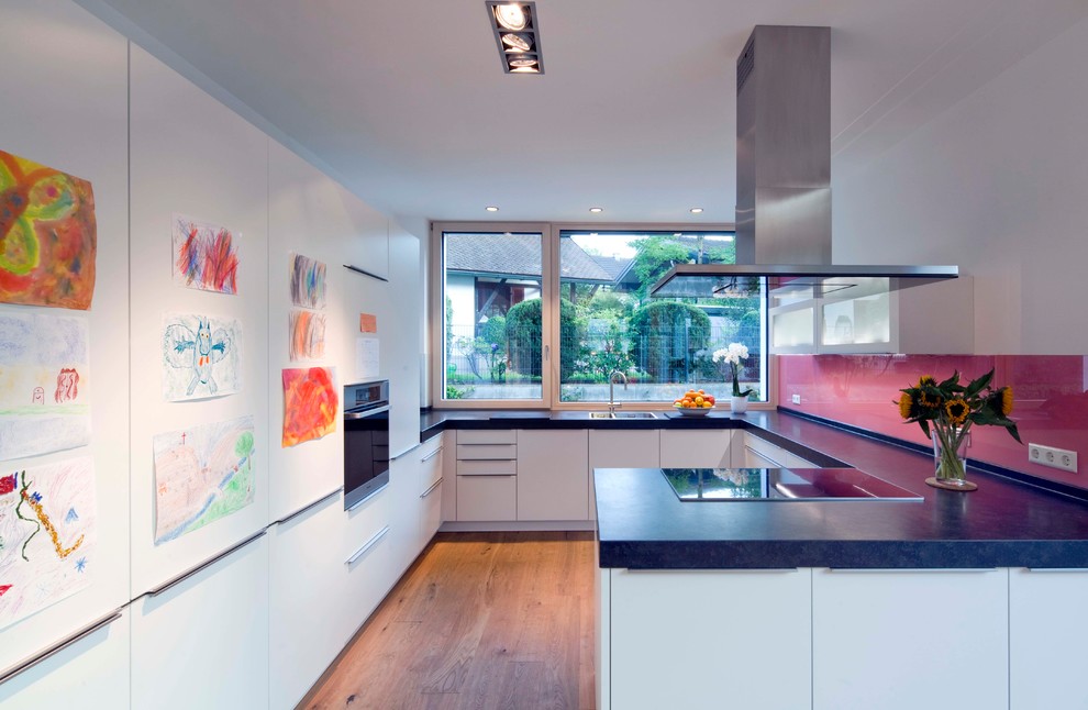 This is an example of a contemporary u-shaped open plan kitchen in Munich with a drop-in sink, flat-panel cabinets, white cabinets, pink splashback, glass sheet splashback, black appliances, medium hardwood floors, a peninsula, brown floor and blue benchtop.