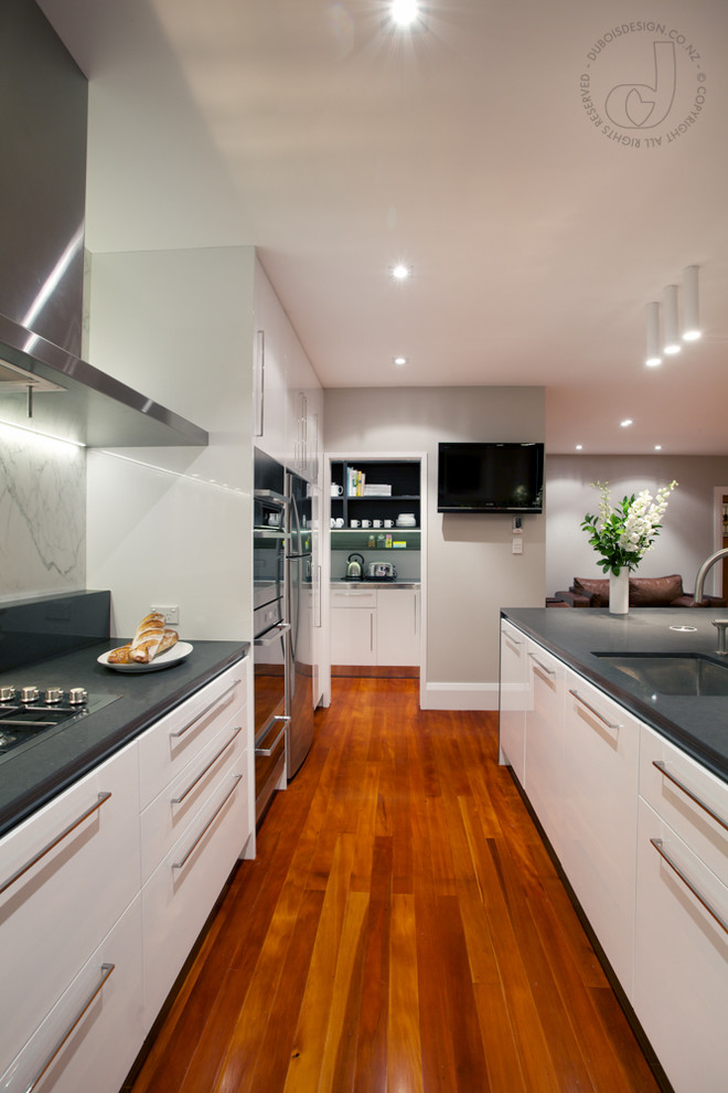 This is an example of a mid-sized contemporary galley kitchen pantry in Auckland with a drop-in sink, glass-front cabinets, white cabinets, quartz benchtops, white splashback, stone slab splashback, stainless steel appliances, medium hardwood floors and with island.