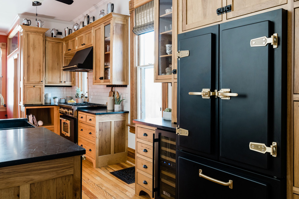 This is an example of a traditional kitchen in Denver with shaker cabinets, medium wood cabinets, white splashback, subway tile splashback, black appliances, medium hardwood floors, with island, brown floor and black benchtop.