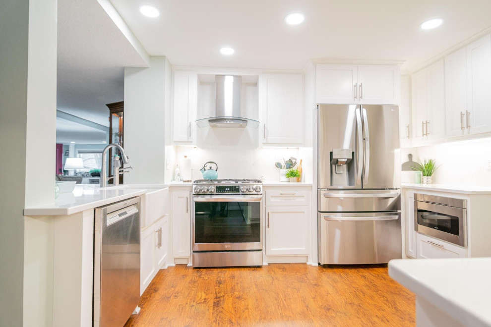 Photo of a small transitional u-shaped separate kitchen in Minneapolis with a farmhouse sink, shaker cabinets, white cabinets, quartz benchtops, white splashback, engineered quartz splashback, stainless steel appliances, vinyl floors, no island, brown floor and white benchtop.