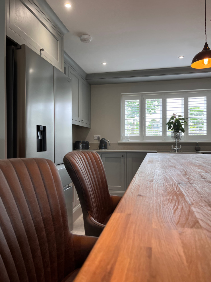 This is an example of a large rustic grey and brown u-shaped kitchen/diner in Essex with a belfast sink, shaker cabinets, grey cabinets, wood worktops, grey splashback, black appliances, an island, brown worktops and feature lighting.