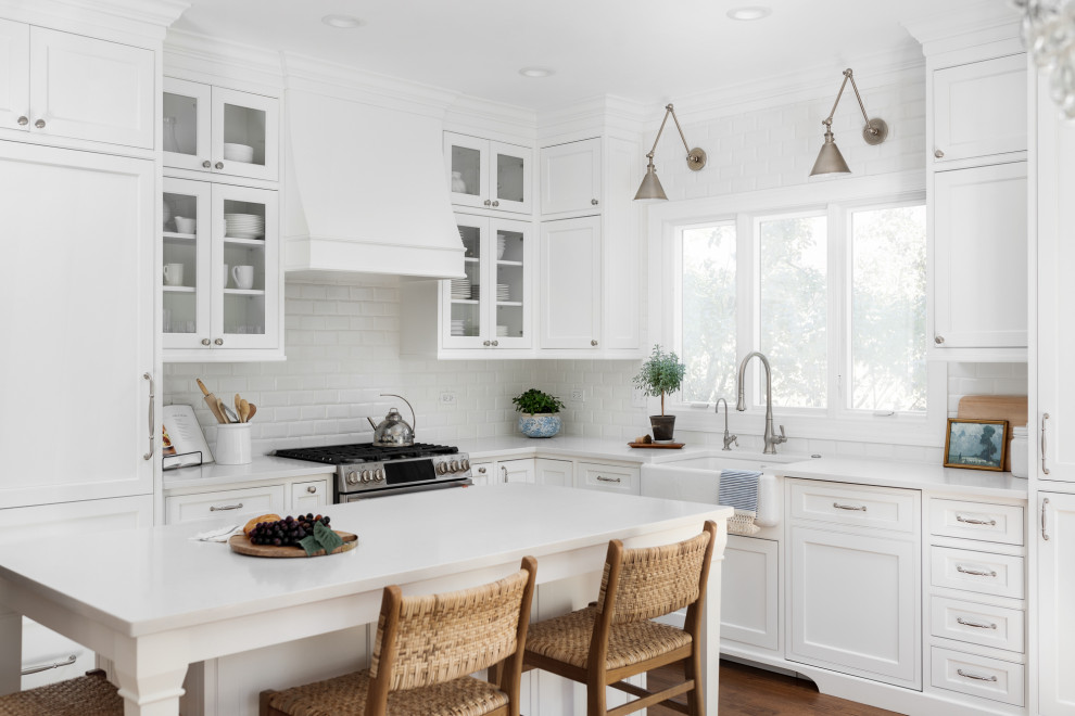Medium sized classic l-shaped kitchen/diner in Chicago with a belfast sink, beaded cabinets, white cabinets, engineered stone countertops, white splashback, ceramic splashback, integrated appliances, medium hardwood flooring, an island, brown floors and white worktops.