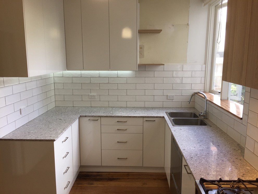 Photo of a small contemporary u-shaped separate kitchen in Melbourne with a double-bowl sink, flat-panel cabinets, white cabinets, granite benchtops, white splashback, stainless steel appliances, medium hardwood floors, ceramic splashback and grey benchtop.