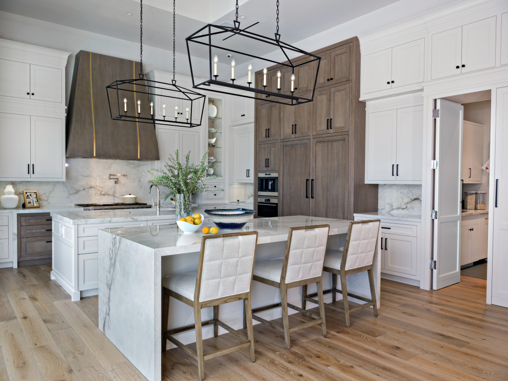 Expansive transitional l-shaped kitchen pantry in Miami with a farmhouse sink, shaker cabinets, white cabinets, solid surface benchtops, white splashback, porcelain splashback, stainless steel appliances, light hardwood floors, multiple islands, brown floor, white benchtop and recessed.