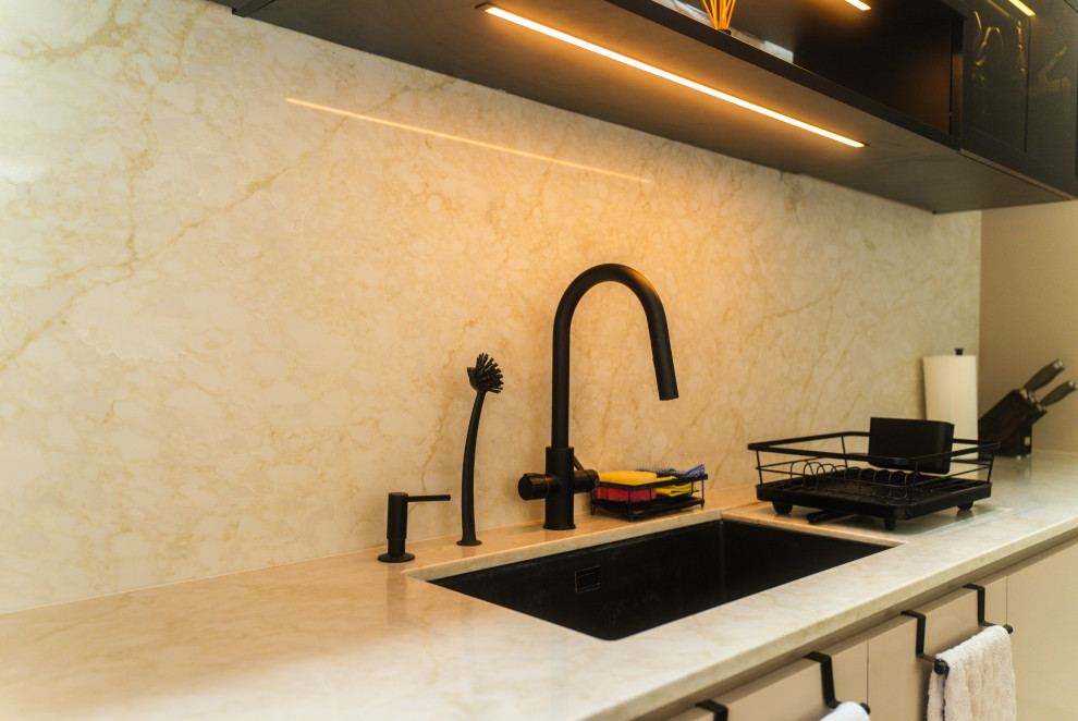 Photo of a medium sized cream and black single-wall open plan kitchen in London with beige cabinets, quartz worktops, beige splashback, engineered quartz splashback, black appliances, porcelain flooring, an island, grey floors, beige worktops and feature lighting.