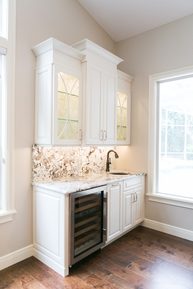 This is an example of a mid-sized transitional single-wall wet bar in Orlando with an undermount sink, white cabinets, granite benchtops, white splashback, medium hardwood floors, stone slab splashback, brown floor, raised-panel cabinets and multi-coloured benchtop.
