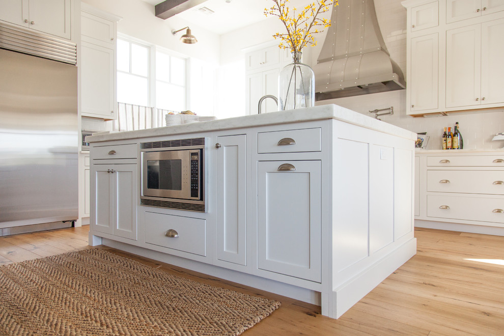 Large transitional l-shaped light wood floor eat-in kitchen photo in Phoenix with a farmhouse sink, white cabinets, white backsplash, stainless steel appliances, an island, stainless steel countertops, shaker cabinets and porcelain backsplash