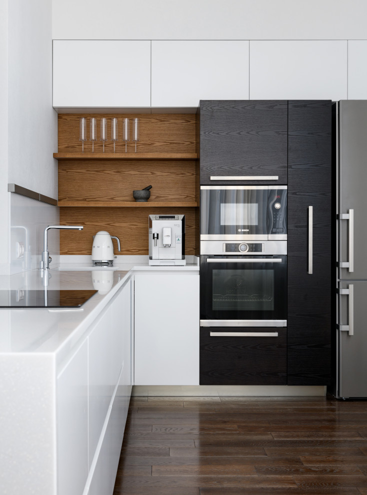This is an example of a mid-sized contemporary l-shaped open plan kitchen in Novosibirsk with an undermount sink, flat-panel cabinets, white cabinets, solid surface benchtops, white splashback, dark hardwood floors, with island, brown floor and white benchtop.