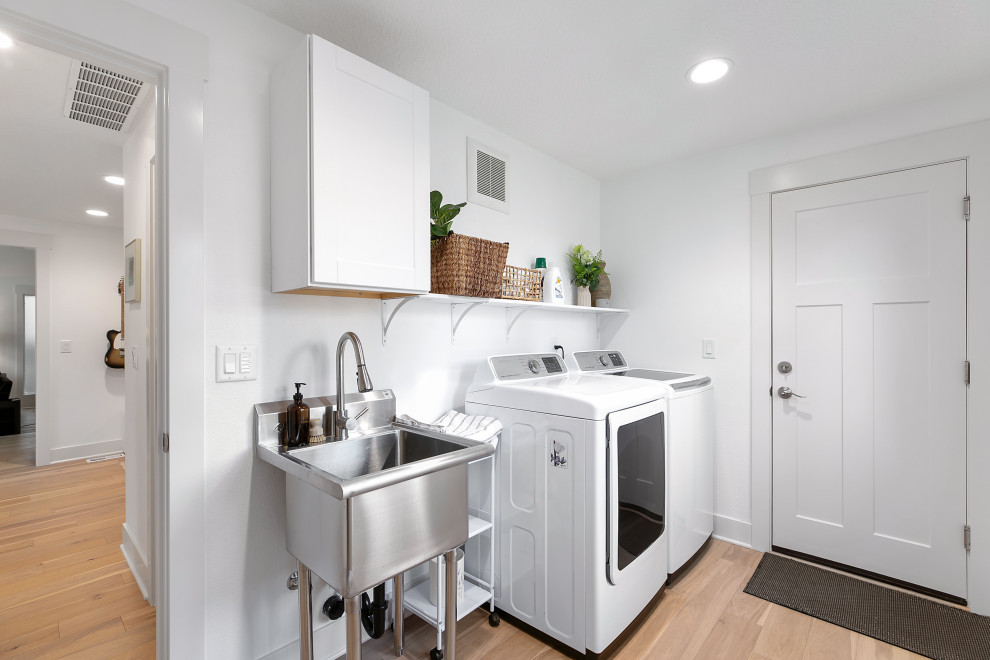 Mid-sized scandinavian galley dedicated laundry room in Portland with an utility sink, shaker cabinets, white cabinets, white walls, light hardwood floors, a side-by-side washer and dryer and brown floor.