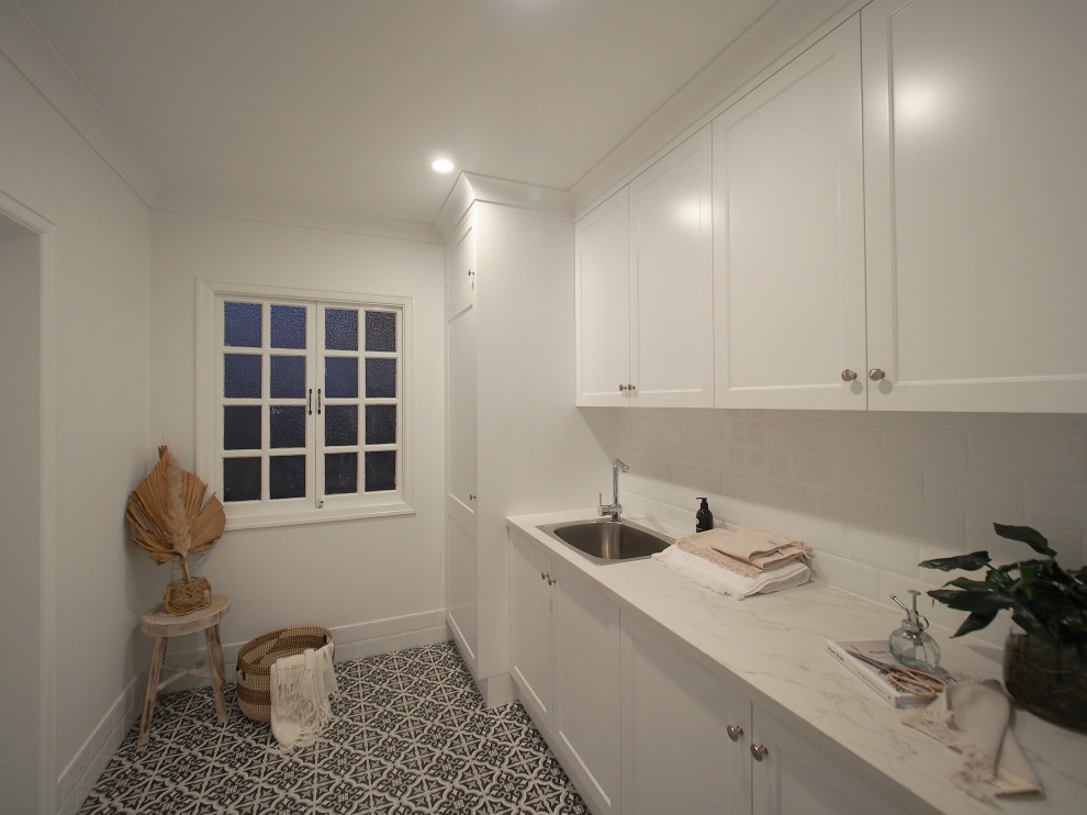 This is an example of a contemporary galley dedicated laundry room in Brisbane with shaker cabinets, white cabinets, marble benchtops, white splashback, ceramic splashback, ceramic floors, a concealed washer and dryer, multi-coloured floor and white benchtop.