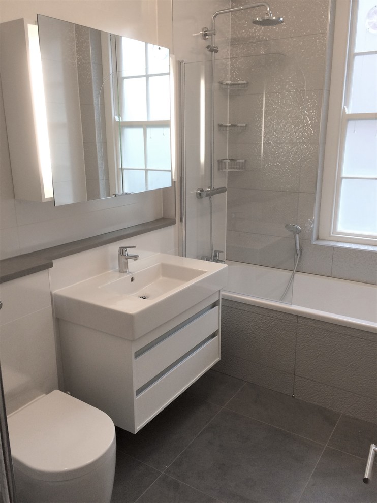 Small contemporary kids bathroom in London with flat-panel cabinets, white cabinets, a drop-in tub, a shower/bathtub combo, a one-piece toilet, white tile, ceramic tile, porcelain floors, a wall-mount sink, quartzite benchtops, grey floor, a hinged shower door and grey benchtops.