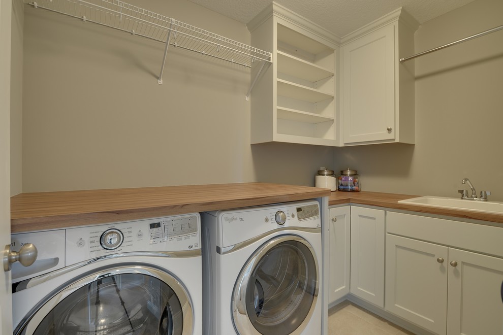Inspiration for a large transitional l-shaped dedicated laundry room in Minneapolis with a drop-in sink, recessed-panel cabinets, white cabinets, wood benchtops, beige walls, ceramic floors and a side-by-side washer and dryer.