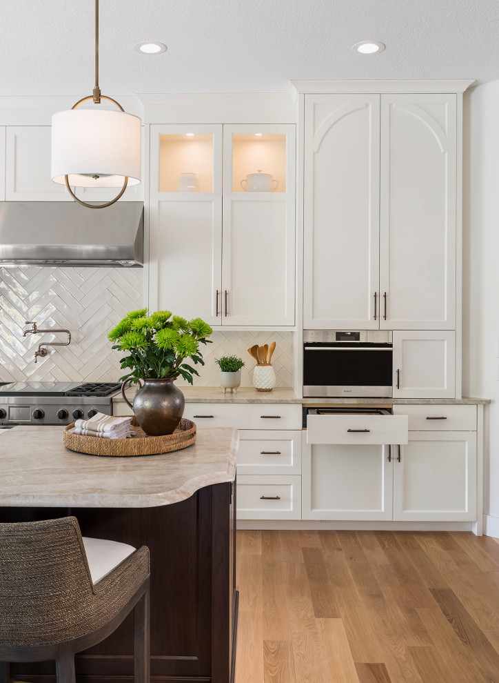 Example of a large transitional single-wall light wood floor and brown floor open concept kitchen design in Minneapolis with a farmhouse sink, recessed-panel cabinets, white cabinets, quartzite countertops, white backsplash, ceramic backsplash, stainless steel appliances, an island and beige countertops