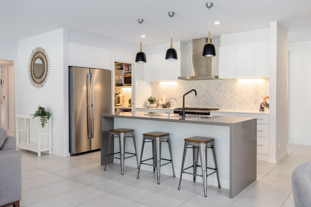 Contemporary l-shaped open plan kitchen in Other with a drop-in sink, flat-panel cabinets, white cabinets, grey splashback, stainless steel appliances, with island, beige floor and white benchtop.