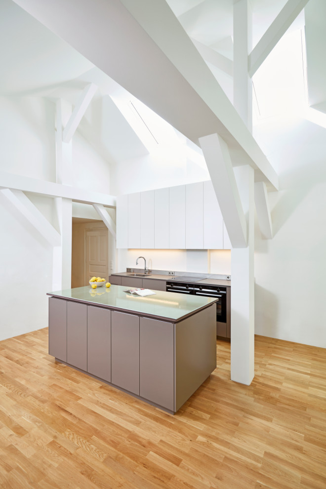 Photo of an expansive contemporary grey and white galley kitchen/diner in Munich with an integrated sink, flat-panel cabinets, grey cabinets, white splashback, black appliances, light hardwood flooring, an island, green worktops and exposed beams.