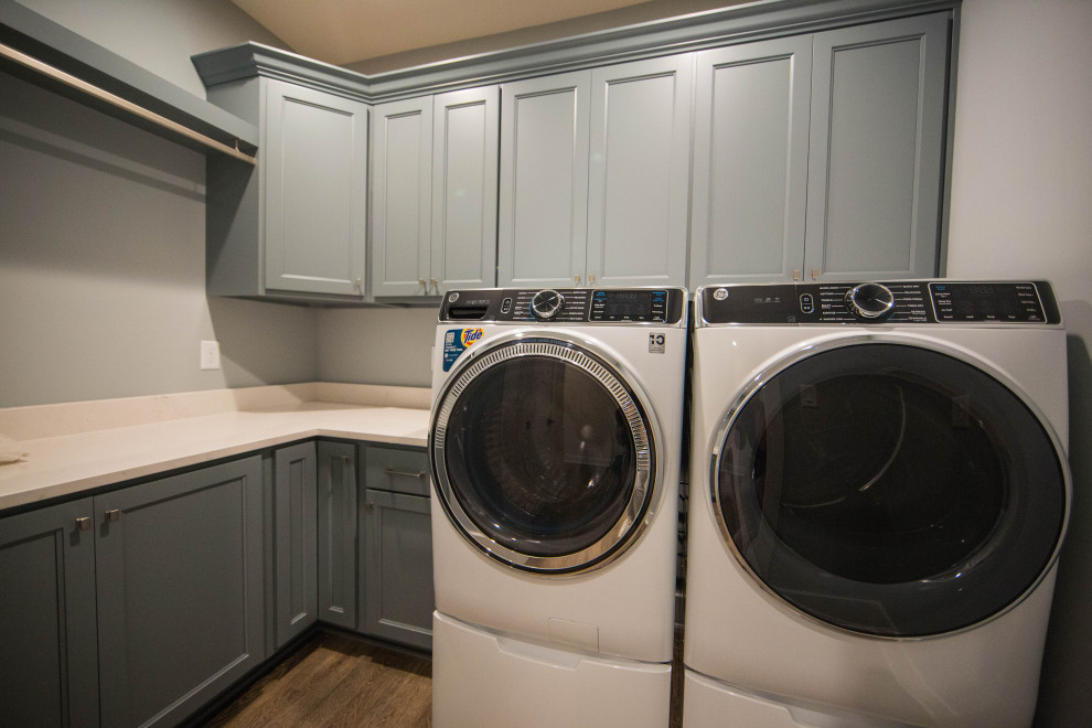 Idéer för mellanstora l-formade vitt tvättstugor enbart för tvätt, med luckor med infälld panel, blå skåp, bänkskiva i kvartsit, grå väggar, laminatgolv, en tvättmaskin och torktumlare bredvid varandra och brunt golv