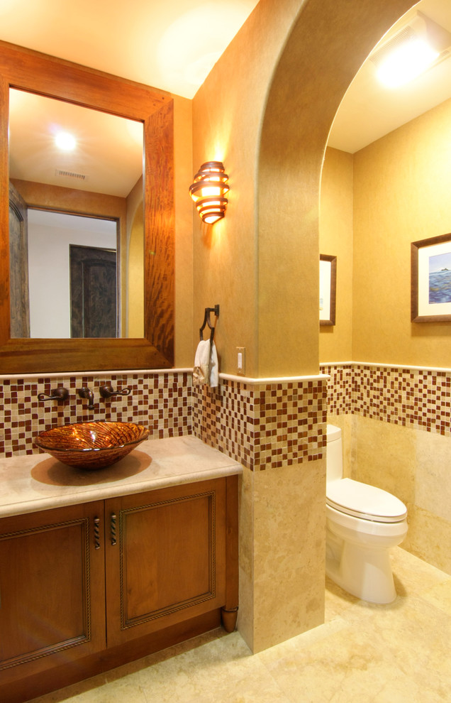 Photo of a mid-sized mediterranean powder room in Los Angeles with recessed-panel cabinets, medium wood cabinets, a one-piece toilet, multi-coloured tile, mosaic tile, beige walls, travertine floors, a vessel sink and limestone benchtops.