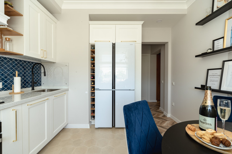 This is an example of a mid-sized traditional l-shaped eat-in kitchen in Other with a single-bowl sink, recessed-panel cabinets, white cabinets, solid surface benchtops, blue splashback, ceramic splashback, black appliances, ceramic floors, beige floor, white benchtop and recessed.