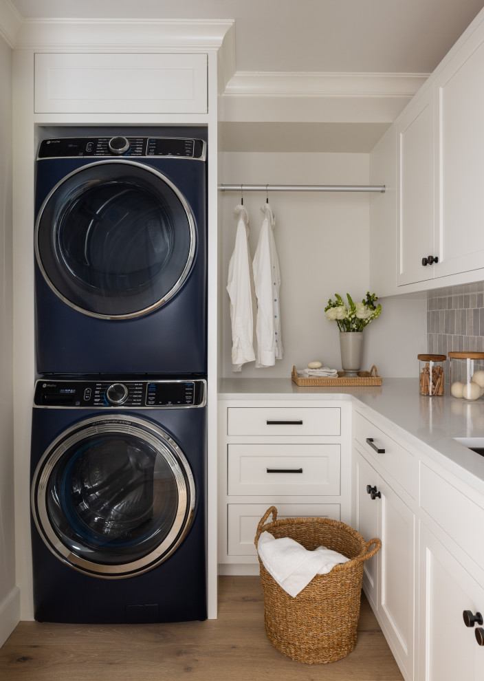 Example of a mid-sized transitional l-shaped dedicated laundry room design in New York with an undermount sink, white cabinets, quartz countertops, green backsplash, ceramic backsplash, a stacked washer/dryer and white countertops