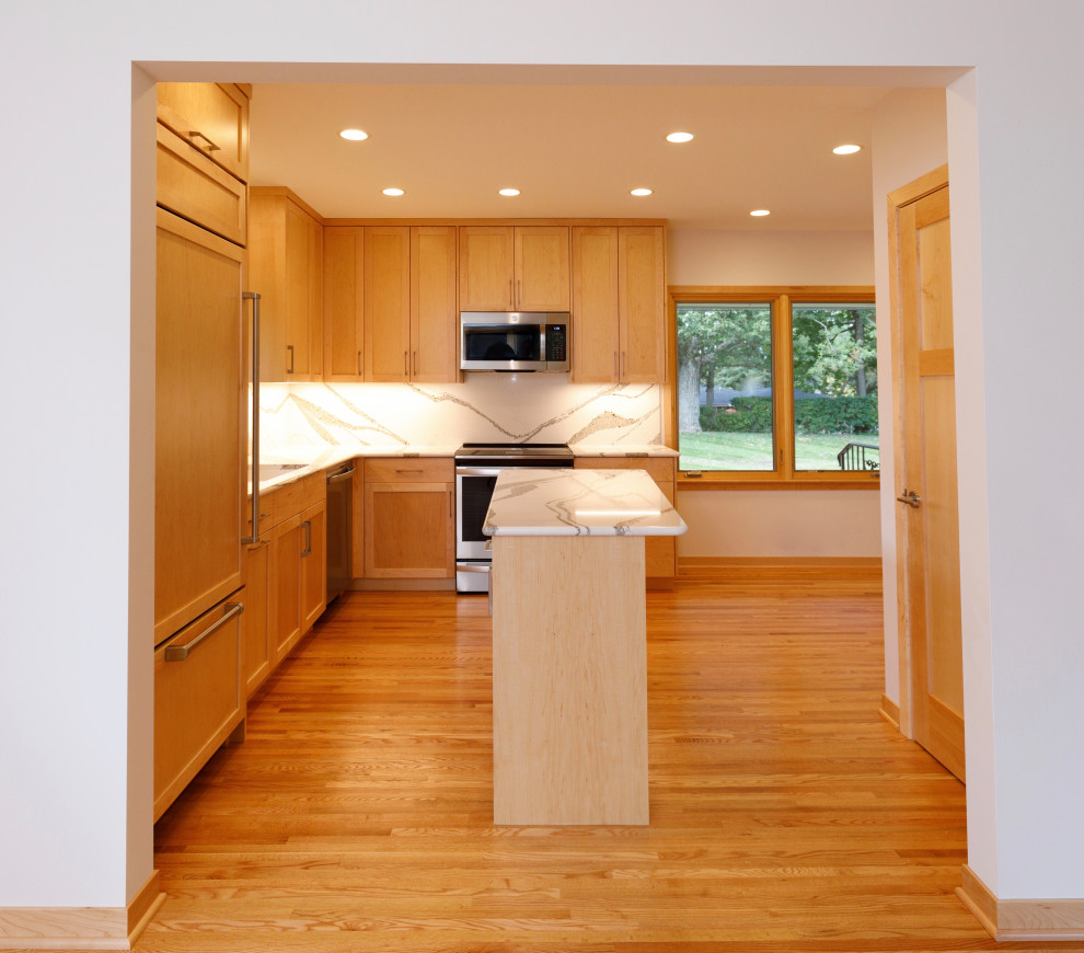 This is an example of a modern l-shaped kitchen in Minneapolis with an undermount sink, shaker cabinets, light wood cabinets, quartz benchtops, white splashback, engineered quartz splashback, stainless steel appliances, light hardwood floors, with island and white benchtop.