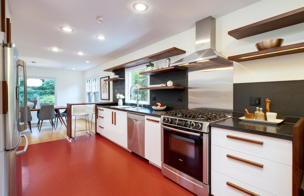 This is an example of a midcentury eat-in kitchen in Portland with flat-panel cabinets, white cabinets, black splashback, stainless steel appliances, a peninsula, red floor and black benchtop.