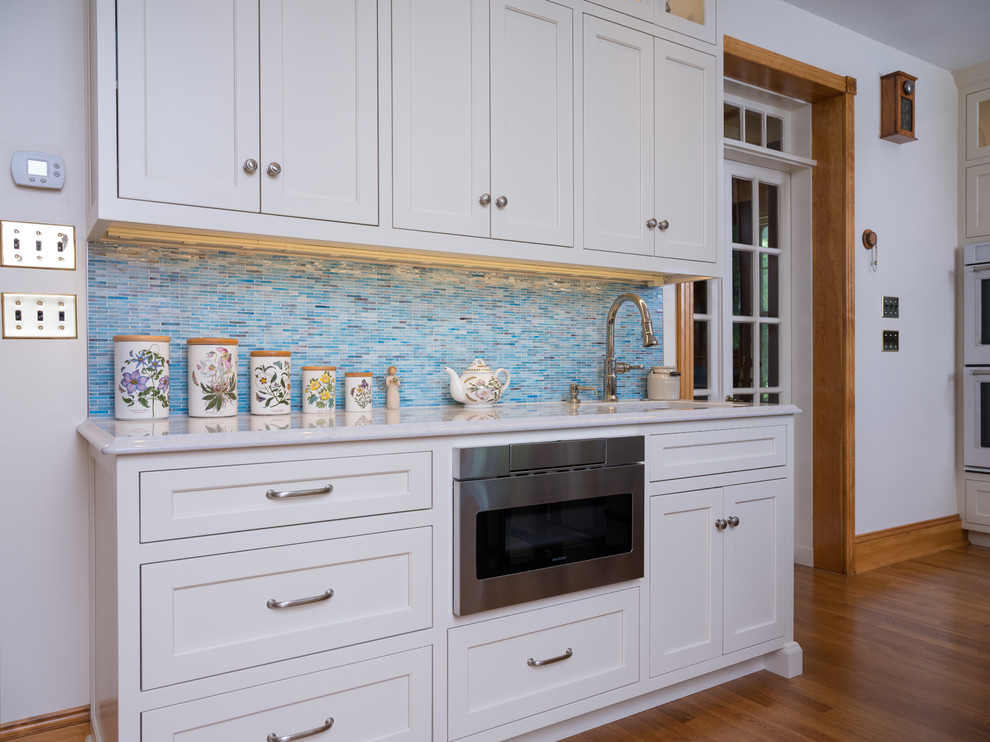 Photo of a large traditional l-shaped open plan kitchen in Philadelphia with an undermount sink, flat-panel cabinets, white cabinets, blue splashback, glass tile splashback, white appliances, light hardwood floors, with island and blue floor.