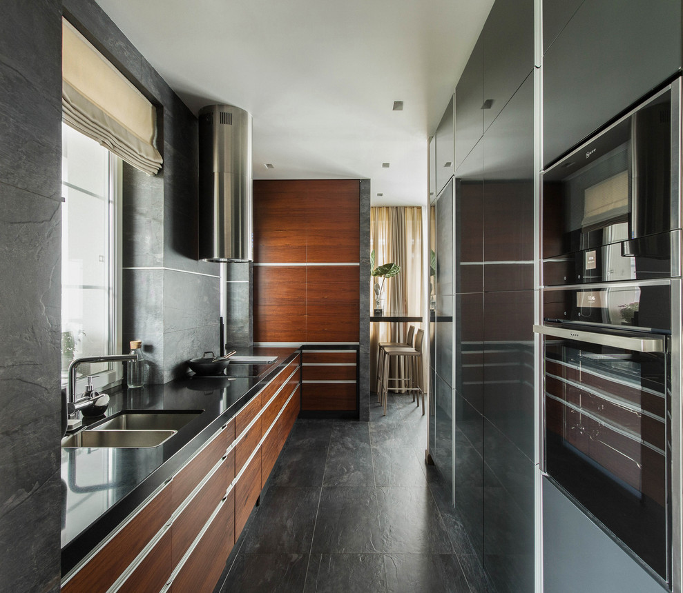 This is an example of a contemporary l-shaped kitchen in Moscow with an undermount sink, flat-panel cabinets, medium wood cabinets, grey splashback, black appliances, grey floor and black benchtop.