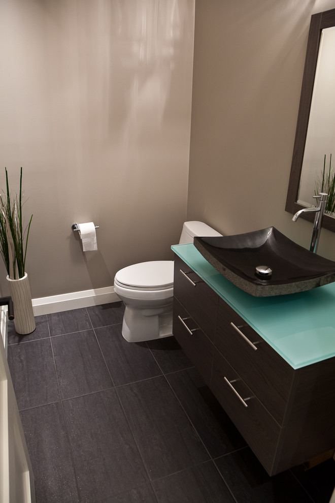 Design ideas for a mid-sized transitional powder room in Calgary with a vessel sink, flat-panel cabinets, dark wood cabinets, glass benchtops, a one-piece toilet, black tile, beige walls and ceramic floors.