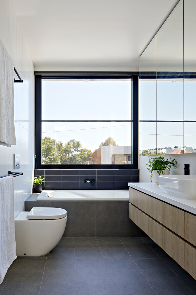 Inspiration for a contemporary bathroom in Melbourne with flat-panel cabinets, light wood cabinets, a drop-in tub, a wall-mount toilet, blue tile, subway tile, white walls, a vessel sink and grey floor.