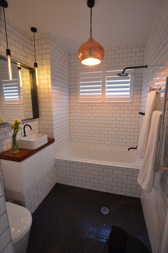 Contemporary bathroom in Sydney with a vessel sink, an alcove tub, a shower/bathtub combo, a one-piece toilet, flat-panel cabinets, white cabinets, wood benchtops, white tile, subway tile, black floor and brown benchtops.