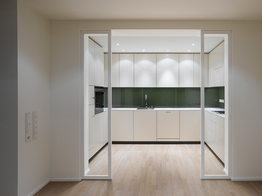 Inspiration for a mid-sized contemporary u-shaped separate kitchen in Stuttgart with flat-panel cabinets, white cabinets, green splashback, glass sheet splashback, light hardwood floors, no island, a drop-in sink and panelled appliances.